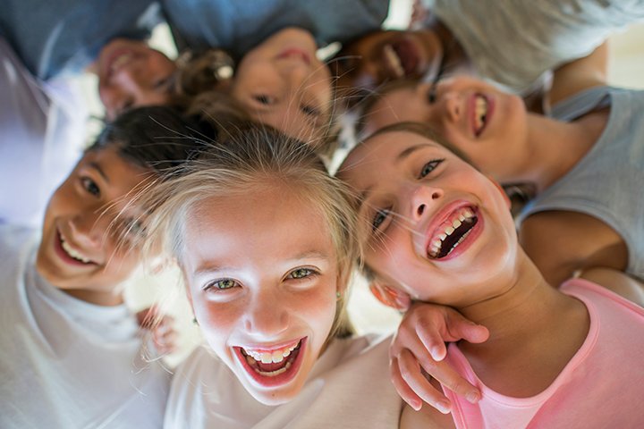 A multi-ethnic group of children are indoors in a health center. They are wearing casual clothing and running shoes. They are embracing and laughing downwards at the camera.